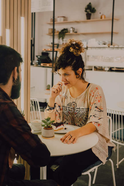 Радісна випадкова жінка їсть торт з виделкою, сидячи за столом у кафетерії, дивлячись на хлопця — стокове фото