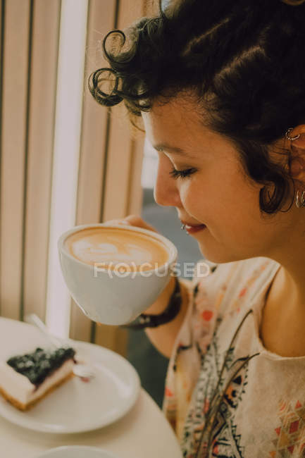 Side view of happy smiling woman smelling mug while sitting in modern coffee shop — Stock Photo