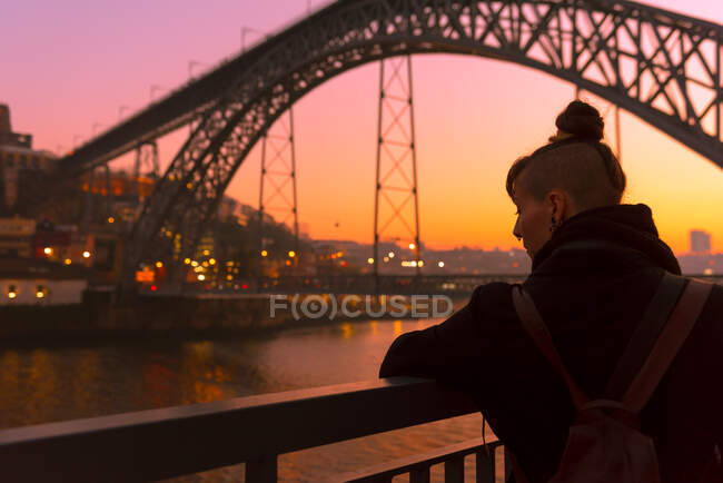 Rückansicht einer Touristin, die in der Nähe des Stadtdamms in der Nähe einer Brücke steht und bei Sonnenuntergang in Porto, Portugal, wegschaut — Stockfoto