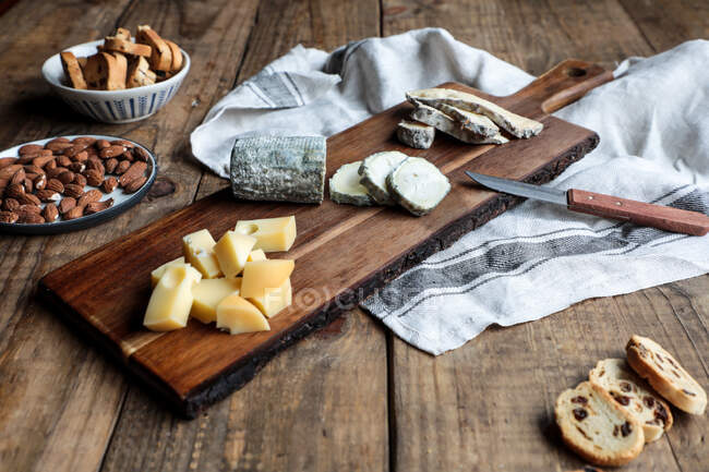 Süße Croutons mit Rosinen und Teller mit Mandeln auf Holztisch neben Brett mit verschiedenen geschnittenen Käse platziert — Stockfoto