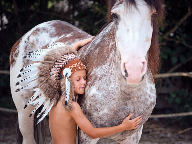 Tranquil child with closed eyes wearing traditional Indian war bonnet bonding with horse stallion on blurred background — Stock Photo