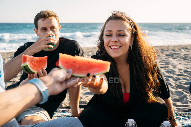 Smiling woman gives a piece of watermelon to her friend with her friend who drinks orange juice on the beach — Stock Photo