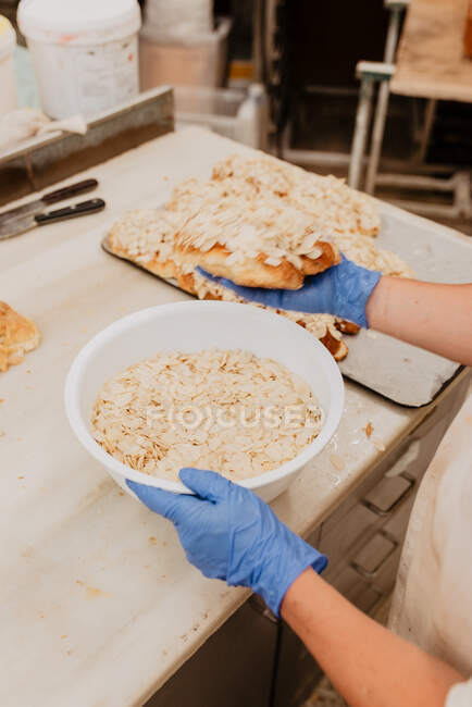 Сверху анонимный кондитер в латексных перчатках держит миску свежих миндальных хлопьев и готовит вкусные кондитерские во время работы в пекарне — стоковое фото