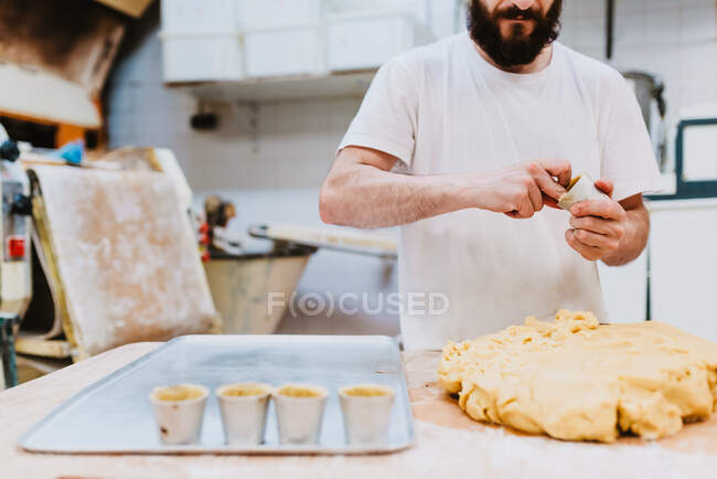 Crop bearded man in white t-shirt putting fresh dough into cups while making pastry in kitchen of bakery — Stock Photo