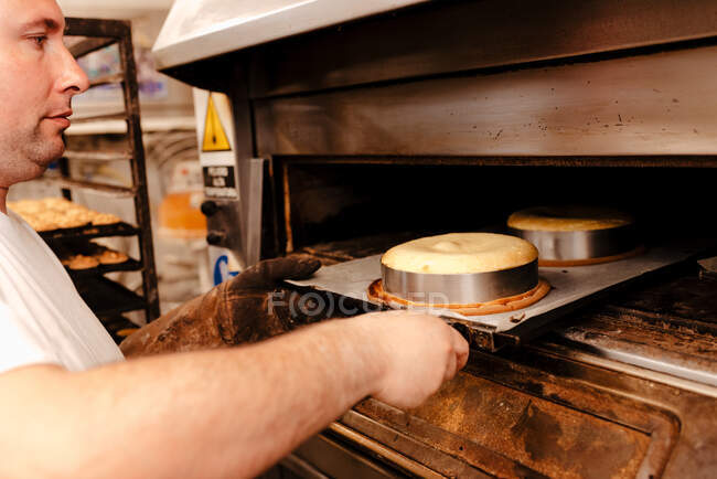 Cultive o homem adulto em bandeja uniforme colocando com bolos crus no forno quente enquanto trabalhava na padaria — Fotografia de Stock
