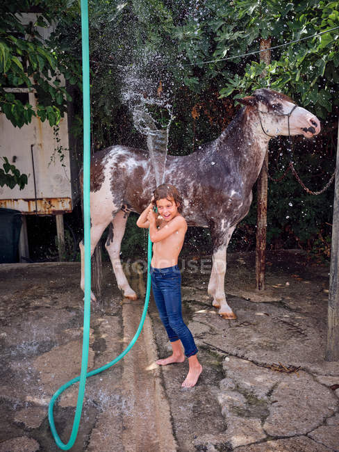 Barefoot boy hosing down stallion with fresh water on farm terrace — Stock Photo