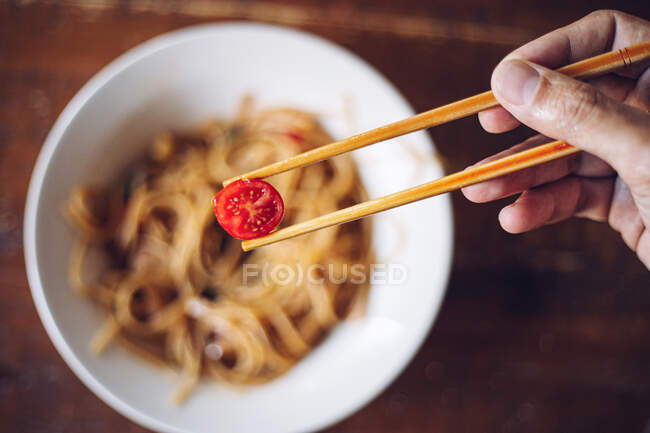 From above soft focus of crop person holding cherry tomato by wooden chopsticks while enjoying noodles on blurred background — Stock Photo