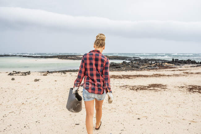 Позитивна хіпстерка в повсякденному одязі і рукавичках, що збирають сміття в сумку, присідаючи на покинутому пляжі — стокове фото