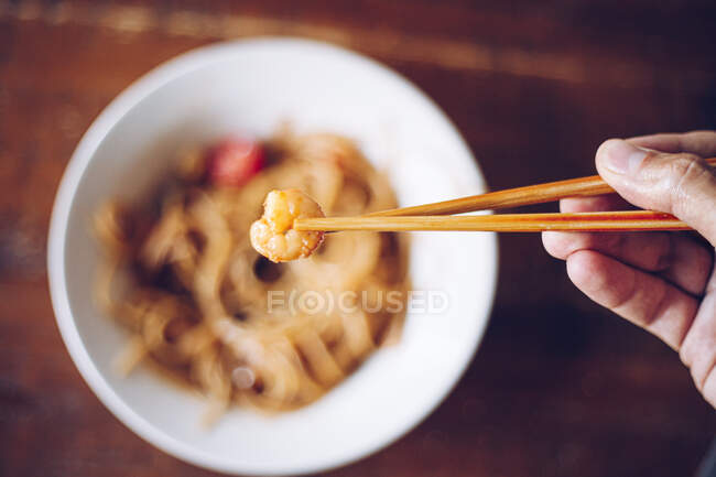 From above soft focus of crop person holding shrimp by wooden chopsticks while enjoying noodles on blurred background — Stock Photo