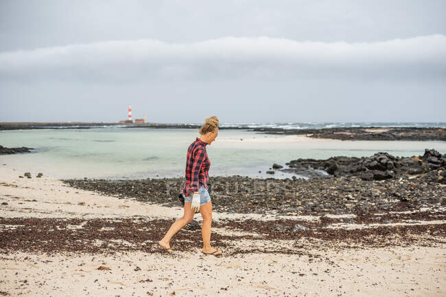 Позитивна хіпстерка в повсякденному одязі і рукавичках, що збирають сміття в сумку, присідаючи на покинутому пляжі — стокове фото