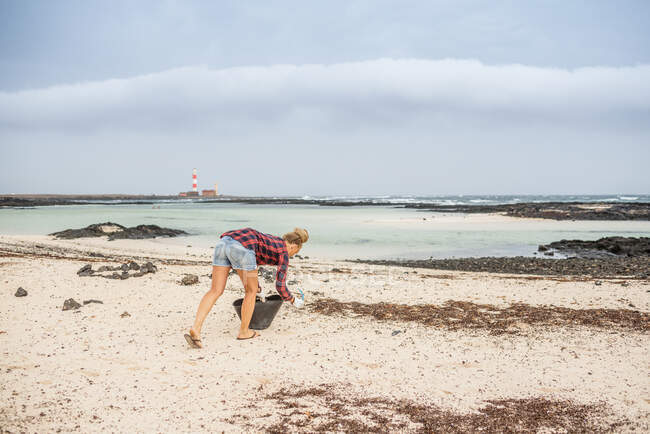 Mujer hipster positiva en ropa casual y guantes recogiendo basura en la bolsa mientras se agacha en la playa desierta - foto de stock