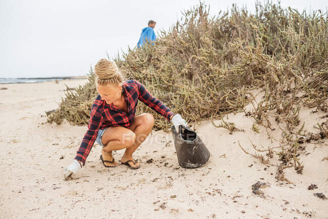 Mulher hipster positivo em roupas casuais e luvas coletando lixo em saco enquanto agachamento na praia deserta — Fotografia de Stock