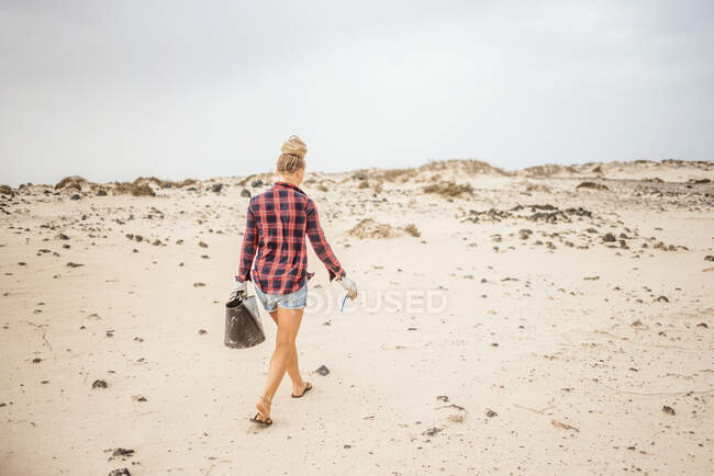 Позитивная хипстерша в повседневной одежде и перчатках собирает мусор в мешок, сидя на пустынном пляже — стоковое фото