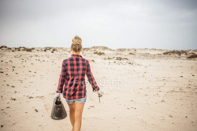Позитивная хипстерша в повседневной одежде и перчатках собирает мусор в мешок, сидя на пустынном пляже — стоковое фото
