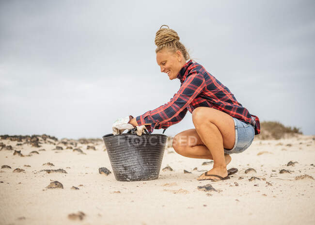 Mujer hipster positiva en ropa casual y guantes recogiendo basura en la bolsa mientras se agacha en la playa desierta - foto de stock
