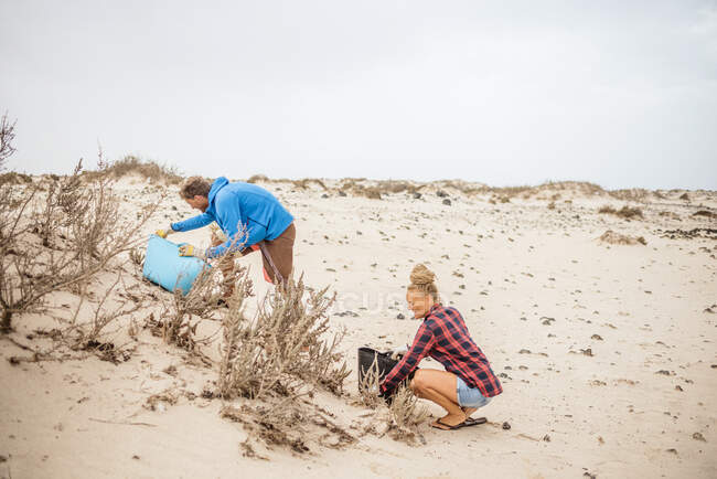 Mulher hipster positivo e homem em roupas casuais e luvas coletando lixo em saco enquanto agachamento na praia deserta — Fotografia de Stock