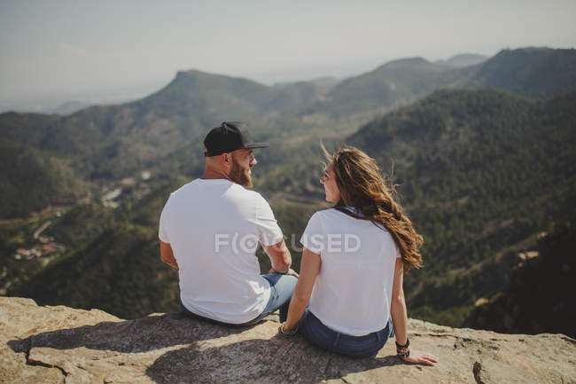 Desde atrás, una pareja romántica y relajada en un equipo que combina la vista mientras se sienta al borde del alto acantilado bajo la luz del sol. - foto de stock
