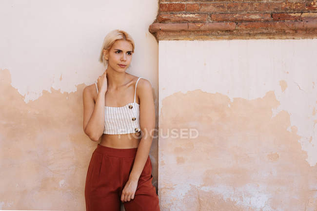 Молода жінка в модному верхній частині і штани, дивлячись далеко, стоячи навпроти міцної будівельної стіни на вулиці стародавнього міста — стокове фото