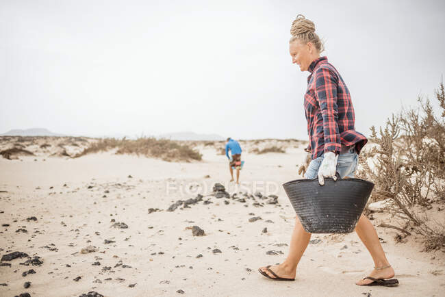 Positivo hipster donna in abiti casual e guanti raccolta spazzatura in borsa mentre accovacciato sulla spiaggia deserta — Foto stock