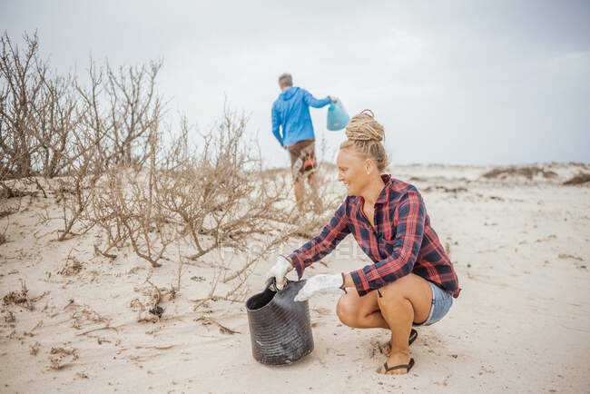 Mujer y hombre hipster positivo en ropa casual y guantes recogiendo basura en la bolsa mientras se agacha en la playa desierta - foto de stock