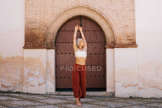 Attractive young woman in stylish outfit dancing with closing eyes against ancient building with shabby gate on street of old town — Stock Photo
