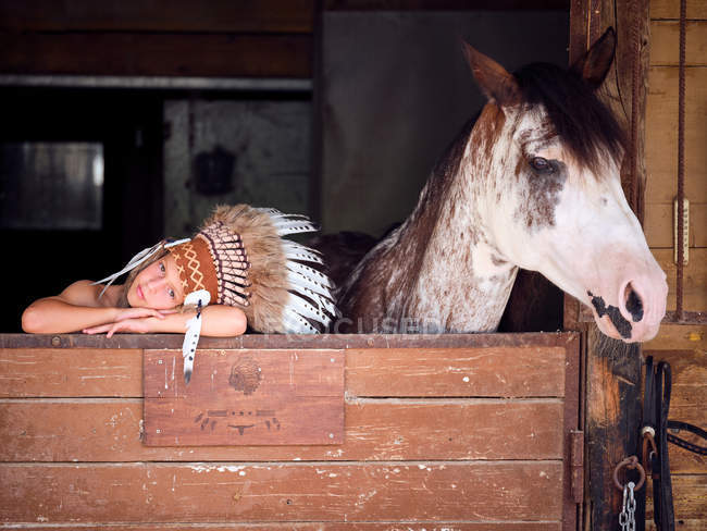 Niño encantado en sombrero de plumas india auténtica apoyada en valla de madera en el establo con caballo en el rancho y mirando a la cámara - foto de stock