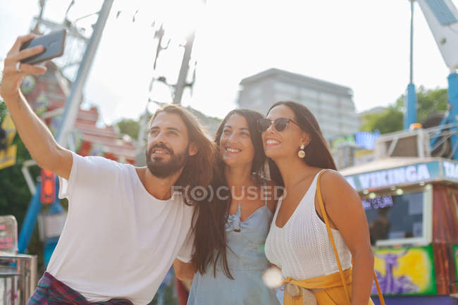 Gente bronceada sonriente alegre tomando fotos en el teléfono inteligente mientras está de pie junto a la atracción en el carnaval - foto de stock
