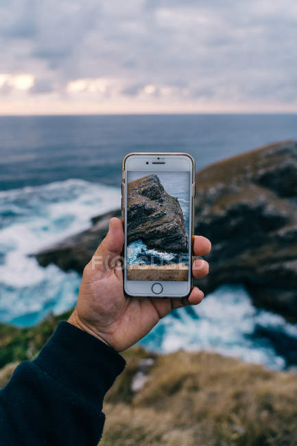 Imagem cortada de viajante segurando smartphone com imagem de penhasco majestoso em ondas oceânicas em pé à beira-mar crepúsculo — Fotografia de Stock