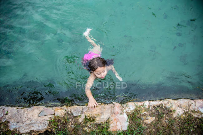 Von oben schwimmt kleines Mädchen zur Steinumrandung im warmen Wasser des Außenpools, während es sich im Wellnessbereich entspannt — Stockfoto