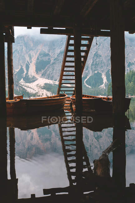Barcos de madera amarrados en el muelle cerca de escaleras en el lago con aguas cristalinas tranquilas sobre un hermoso fondo de montaña brumosa y bosque denso verde en Dolomitas - foto de stock
