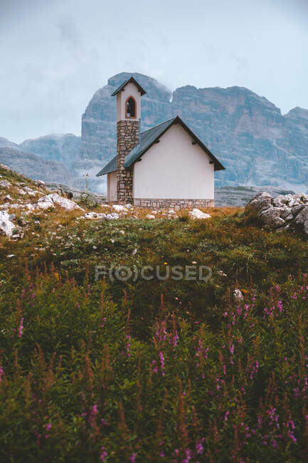 Белое и серое здание на скалистом холме с зеленой толстой травой против красивых туманных гор в Доломитовых Альпах во время пасмурной погоды — стоковое фото