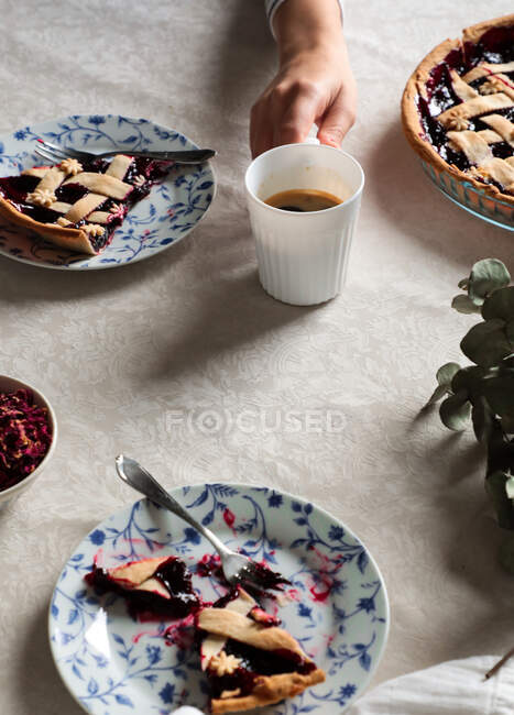 De arriba persona de la cosecha sentado en la mesa cubierta con café servido y delicioso pastel de bayas al horno sobre fondo blanco - foto de stock