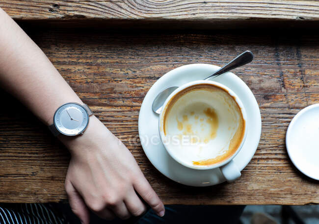 From above crop person with watch on hand sitting at wooden table with empty cup in cafeteria — Stock Photo