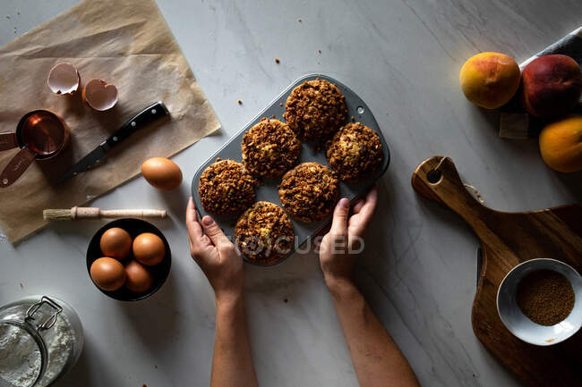 De dessus personne de culture tenant plat de cuisson avec cupcakes faits maison sur une table en bois avec des œufs arrangés avec des pêches et de la farine — Photo de stock
