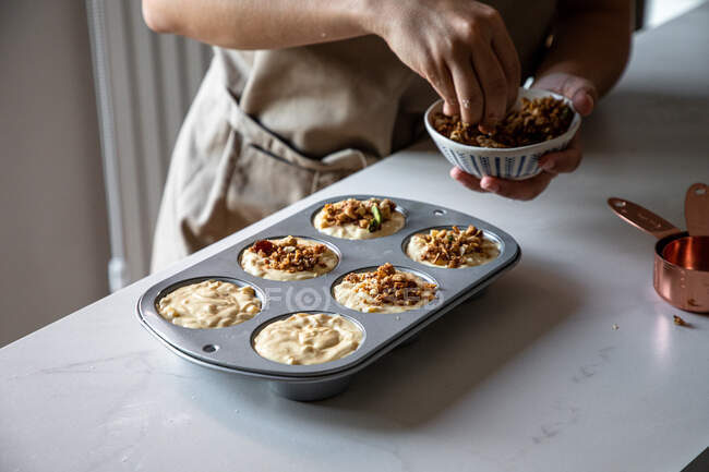 Crop person in apron sprinkling raw homemade cupcakes in baking dish with crushed nuts in kitchen — Stock Photo