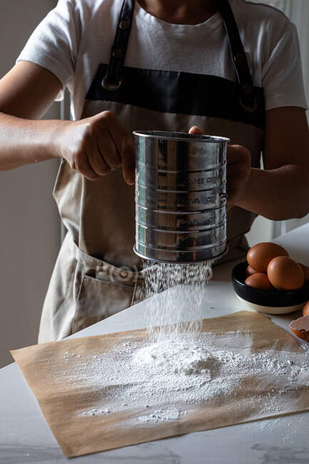 Crop person in grembiule in piedi a tavola setacciando farina su carta da forno con setaccio inox per fare l'impasto — Foto stock