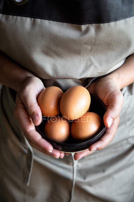 Сверху кукуруза в сером фартуке стоит со свежими куриными яйцами в руках для приготовления пищи — стоковое фото