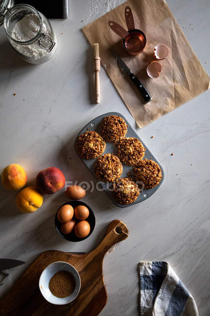 From above composition of delicious cupcakes with walnuts in baking dish eggs peaches flour and parchment paper arranged on white surface — Stock Photo