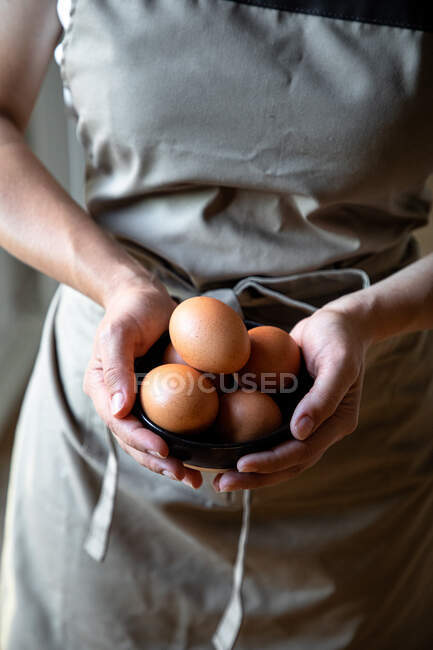 Сверху кукуруза в сером фартуке стоит со свежими куриными яйцами в руках для приготовления пищи — стоковое фото
