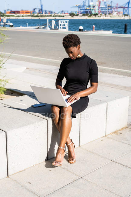 Mujer afroamericana concentrada en elegante vestido negro con portátil en el pavimento - foto de stock
