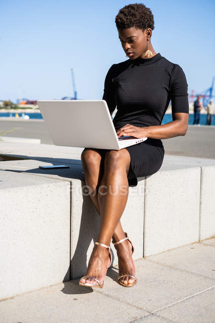 Концентрированная афроамериканка в элегантном черном платье с ноутбуком на тротуаре — стоковое фото