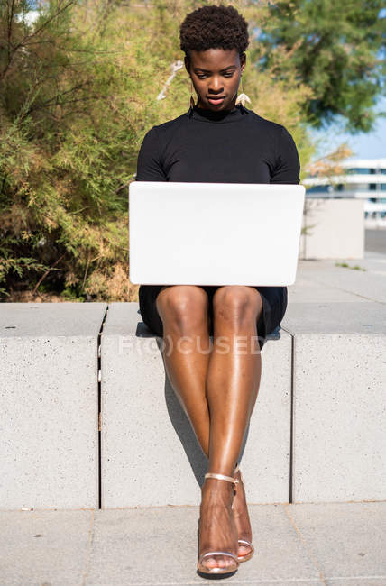Femme afro-américaine concentrée dans une élégante robe noire en utilisant un ordinateur portable sur le trottoir — Photo de stock