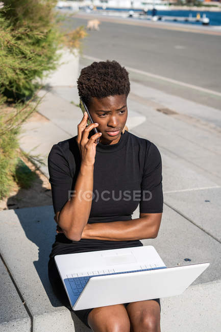 Mujer afroamericana decepcionada en vestido negro usando el ordenador portátil y hablando en el teléfono inteligente en la calle - foto de stock