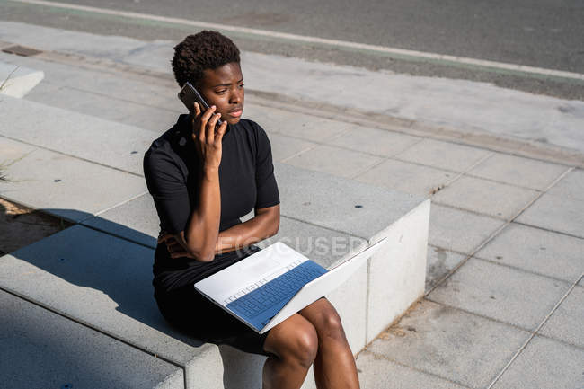 Mulher afro-americana em vestido preto elegante usando laptop e falando no telefone celular na rua — Fotografia de Stock
