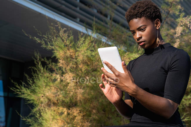 Donna africana in elegante abito nero utilizzando il telefono cellulare mentre seduto sul marciapiede sulla strada — Foto stock