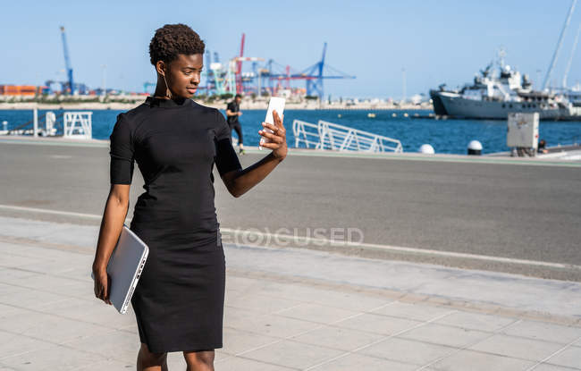 Mujer afroamericana en elegante vestido negro que sostiene el ordenador portátil y el uso de teléfono móvil mientras está de pie en el pavimento - foto de stock