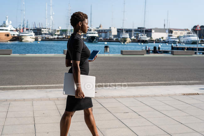 Mulher afro-americana em elegante vestido preto segurando laptop e usando telefone celular enquanto está em pé no pavimento — Fotografia de Stock