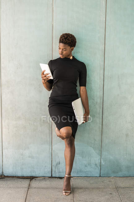 Cool mujer inteligente afroamericana usando un teléfono móvil sosteniendo un ordenador portátil mientras se apoya en la pared de hormigón - foto de stock