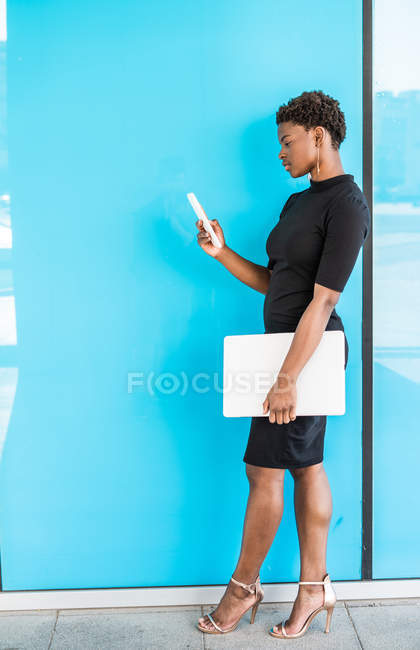 Cool femme intelligente afro-américaine utilisant un téléphone mobile tenant ordinateur portable tout en se tenant debout sur fond bleu — Photo de stock