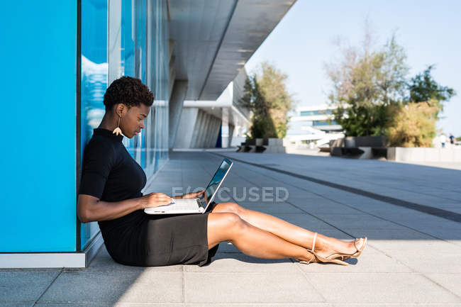 Vue latérale de la femme afro-américaine concentrée dans une élégante robe noire en utilisant un ordinateur portable tout en se relaxant sur le trottoir sur la rue — Photo de stock